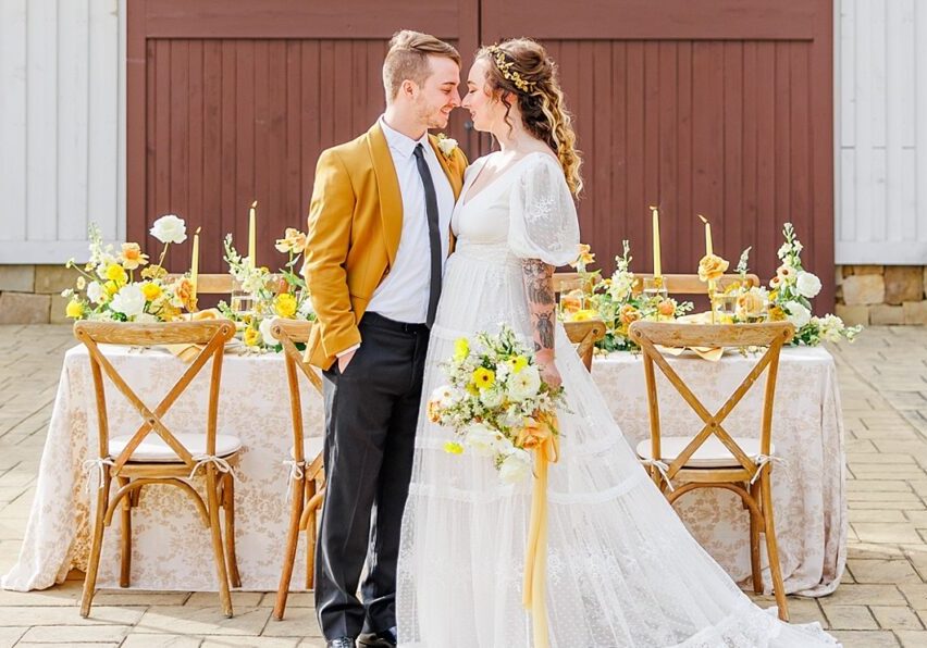 A couple stands in front of a decorated wedding table. The man wears a mustard jacket; the woman is in a white lace dress holding a bouquet. The backdrop features a wooden barn door.