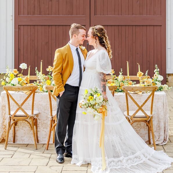 A couple stands in front of a decorated wedding table. The man wears a mustard jacket; the woman is in a white lace dress holding a bouquet. The backdrop features a wooden barn door.