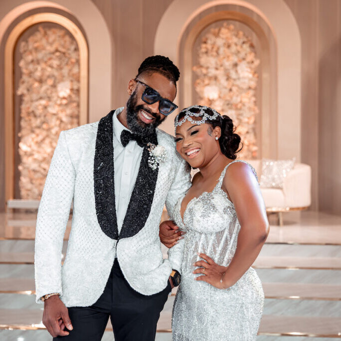 A couple dressed in formal attire poses together. The man is wearing a white tuxedo with black details and sunglasses, while the woman is in a silver gown with matching headpiece. They smile broadly.