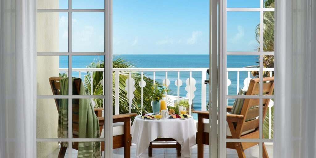 Two wooden chairs and a small table with breakfast items overlook the ocean from a balcony, viewed through open glass doors on a sunny day.