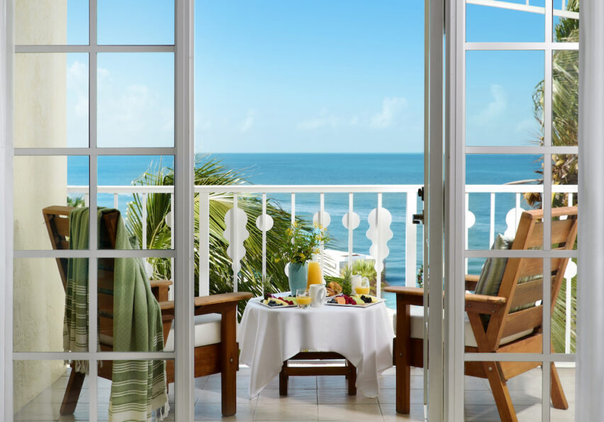 Two wooden chairs and a small table with breakfast items overlook the ocean from a balcony, viewed through open glass doors on a sunny day.