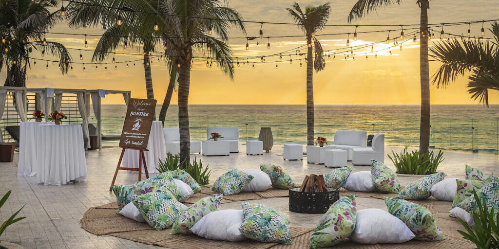 A bonfire on a beach at sunset. The bonfire is surrounded by colorful pillows for seating.