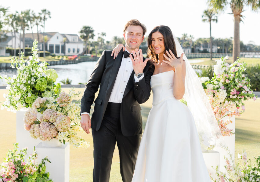 A bride and groom stand outdoors in front of floral arrangements, smiling and showing their wedding rings.