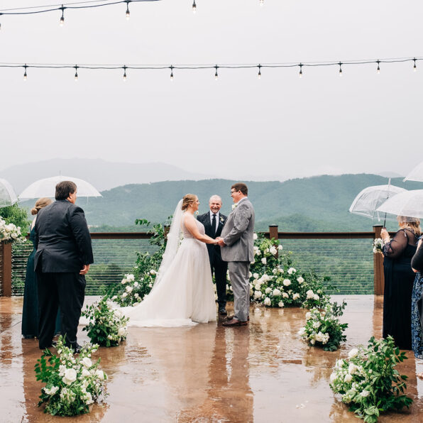 A bride and groom stand at an outdoor wedding ceremony under string lights, holding hands, while guests observe with umbrellas on a rainy day. The background shows a foggy mountain landscape.