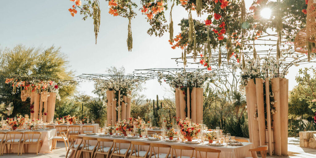 Outdoor wedding reception setup with long tables, floral centerpieces, hanging flowers, and sunlight streaming through greenery.