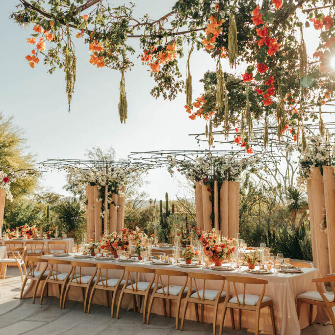 Outdoor wedding reception setup with long tables, floral centerpieces, hanging flowers, and sunlight streaming through greenery.
