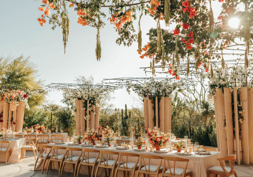 Outdoor wedding reception setup with long tables, floral centerpieces, hanging flowers, and sunlight streaming through greenery.