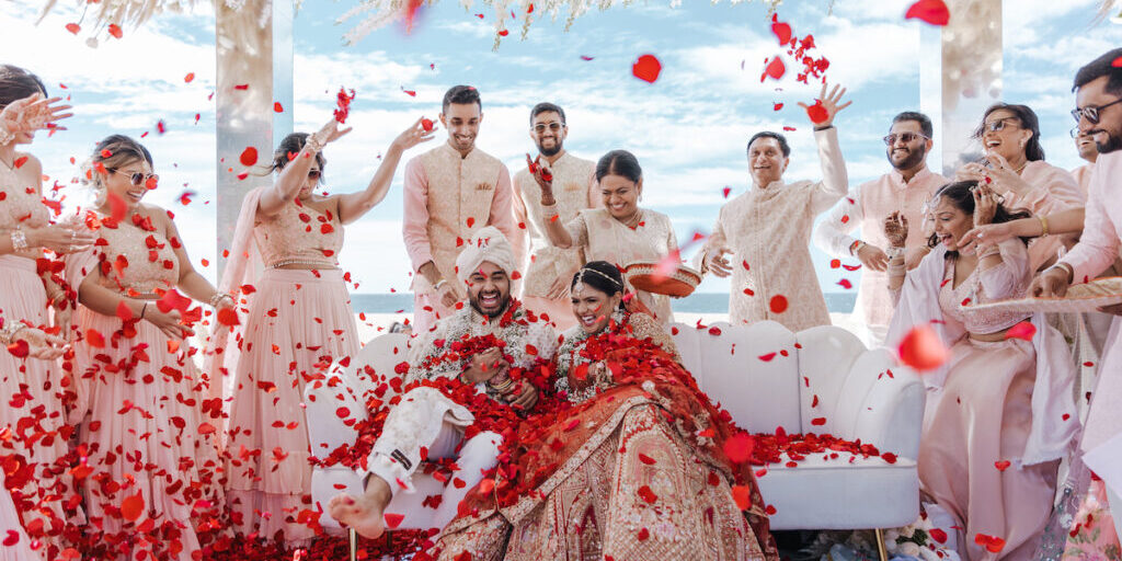 A bride and groom sit surrounded by friends as red rose petals are thrown in the air during a traditional wedding ceremony under a white floral canopy.