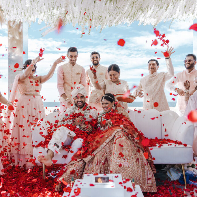A bride and groom sit surrounded by friends as red rose petals are thrown in the air during a traditional wedding ceremony under a white floral canopy.