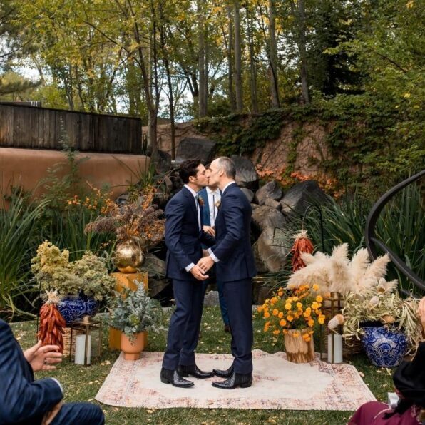 Two men in suits kiss at an outdoor wedding ceremony, standing on a rug surrounded by plants and decorative vases, with greenery and trees in the background.