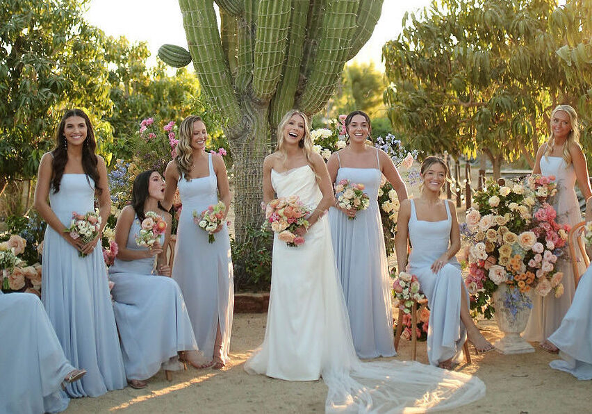 A bride in a white dress stands outdoors with six bridesmaids in light blue dresses, all holding bouquets, near a large cactus and colorful floral arrangements.