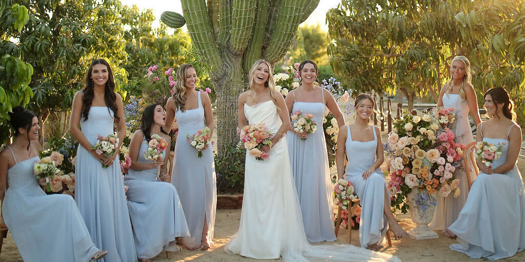 A bride in a white dress stands outdoors with six bridesmaids in light blue dresses, all holding bouquets, near a large cactus and colorful floral arrangements.