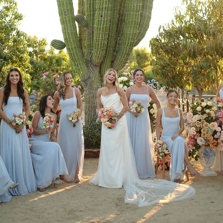 A bride in a white dress stands outdoors with six bridesmaids in light blue dresses, all holding bouquets, near a large cactus and colorful floral arrangements.
