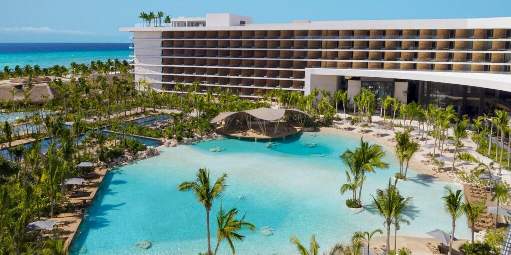 A modern beachfront resort with a large lagoon-style pool, palm trees, and a multi-story hotel building under a clear blue sky.