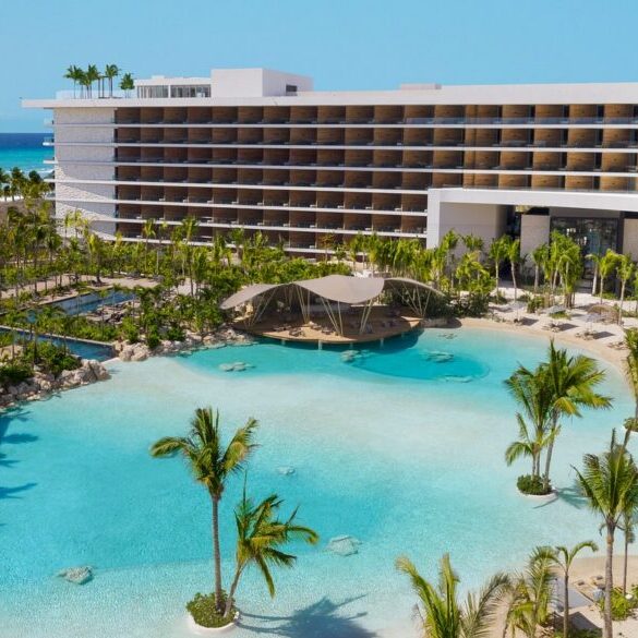 A modern beachfront resort with a large lagoon-style pool, palm trees, and a multi-story hotel building under a clear blue sky.
