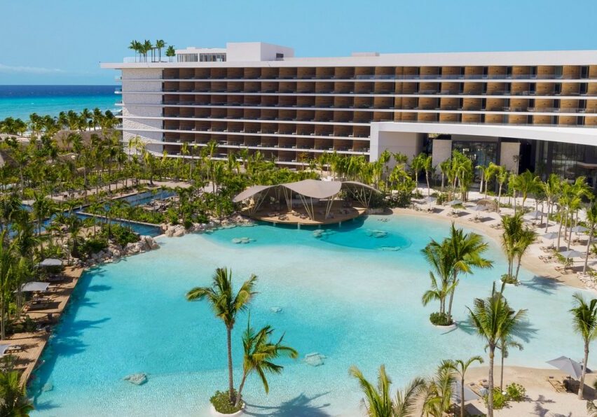 A modern beachfront resort with a large lagoon-style pool, palm trees, and a multi-story hotel building under a clear blue sky.