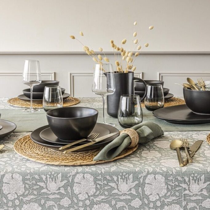 A dining table is set with black dinnerware, gold cutlery, and glasses on a patterned tablecloth. A black vase with dried flowers is in the center.