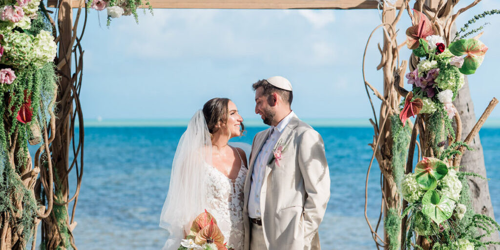 A bride and groom stand under a floral wedding arch on a Islamorada beach, facing each other and smiling, with the ocean in the background.