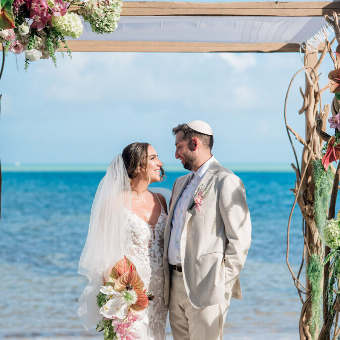 A bride and groom stand under a floral wedding arch on a Islamorada beach, facing each other and smiling, with the ocean in the background.
