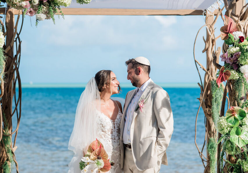 A bride and groom stand under a floral wedding arch on a Islamorada beach, facing each other and smiling, with the ocean in the background.