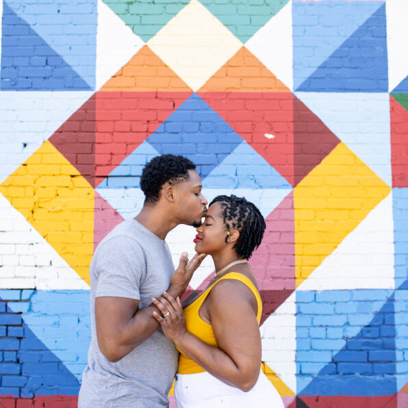 A couple stands in front of a colorful geometric mural. The man kisses the woman's forehead as they hold hands.