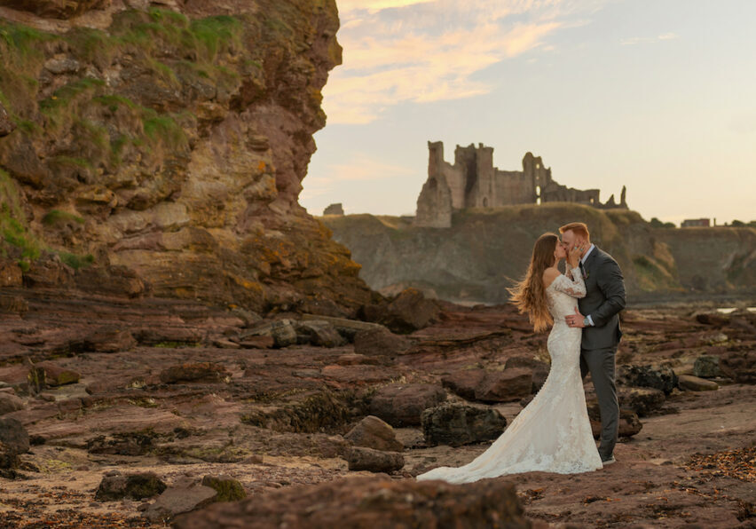 couple pose near ocean with Scottish castle in the background