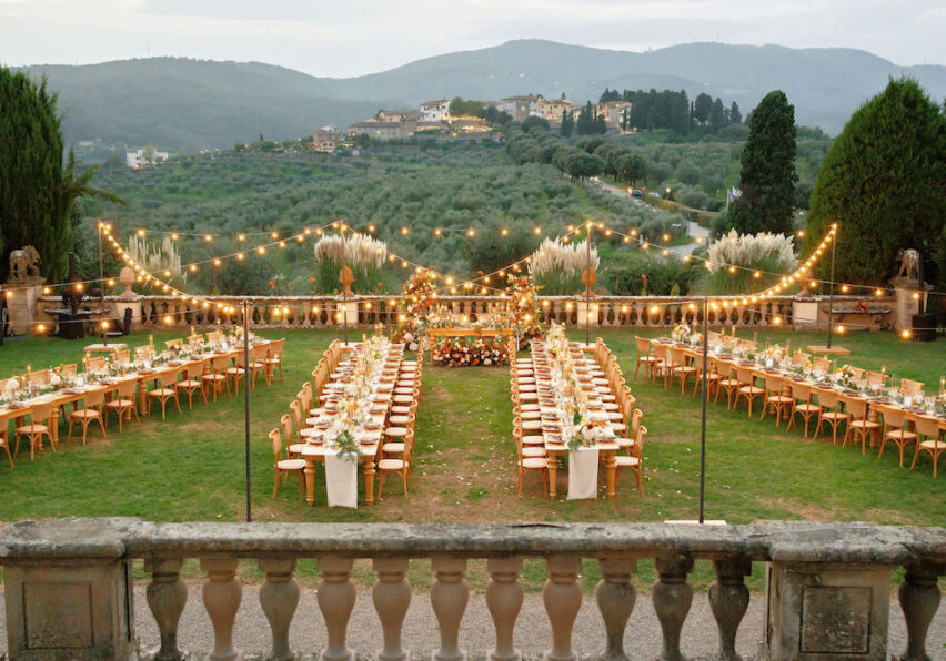 An outdoor formal dining setup with long tables, wooden chairs, and string lights, arranged on a lawn with hills and buildings visible in the background.