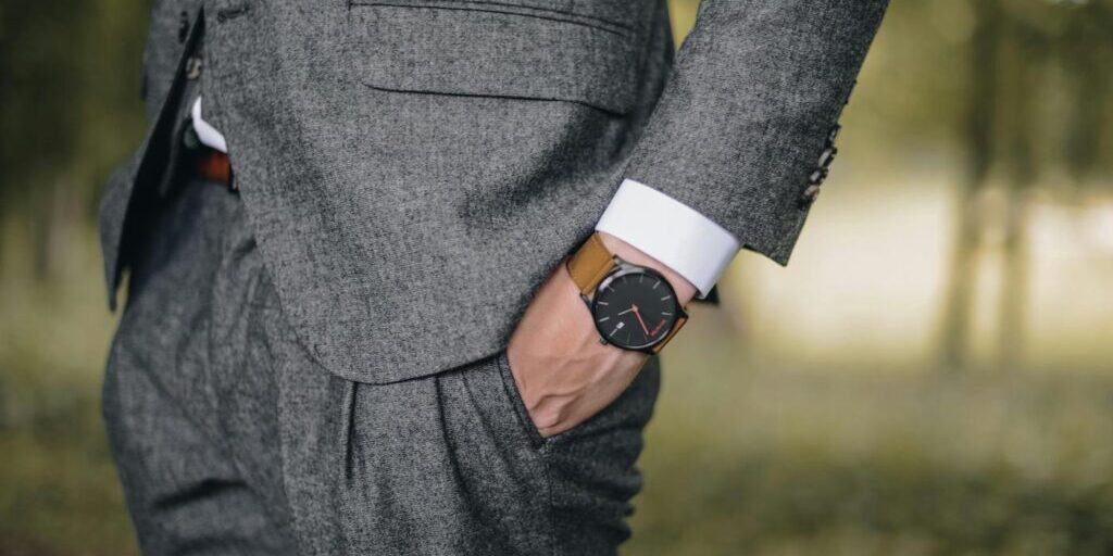 A person wearing a grey suit with their right hand in their pocket, showing a brown leather wristwatch.