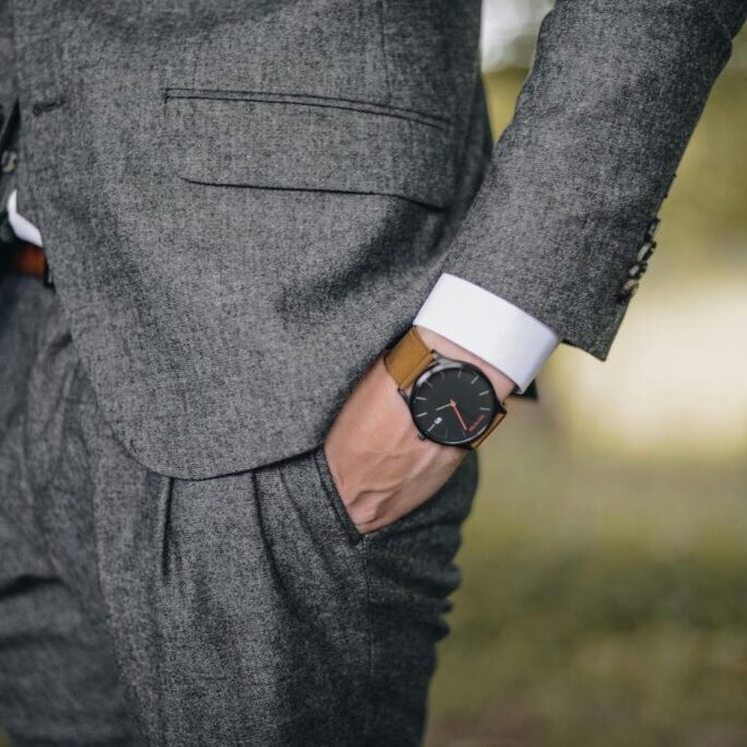 A person wearing a grey suit with their right hand in their pocket, showing a brown leather wristwatch.