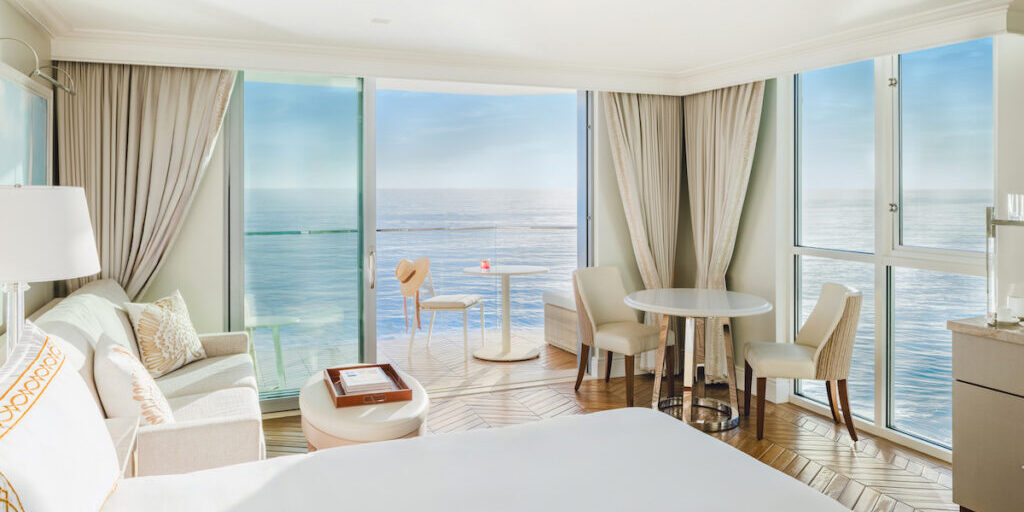 Bright hotel room with large windows, ocean view, bed, sofa, table, chairs, and balcony with a small table and red drink.