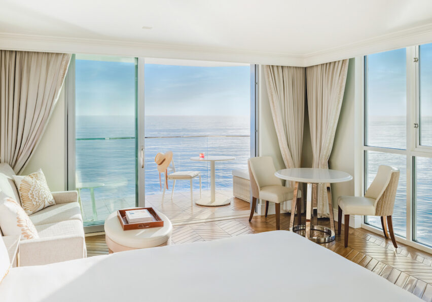 Bright hotel room with large windows, ocean view, bed, sofa, table, chairs, and balcony with a small table and red drink.