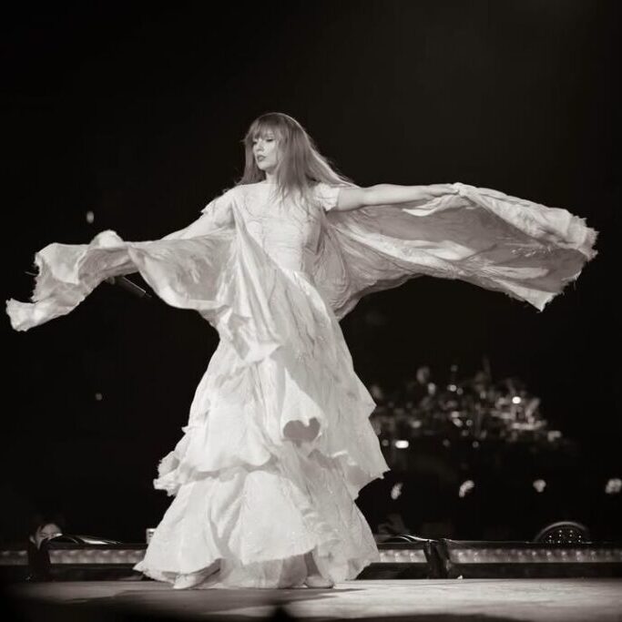 A woman in a flowing dress dances on stage with her arms extended, creating a dramatic, wing-like effect with the fabric. The image is in black and white.