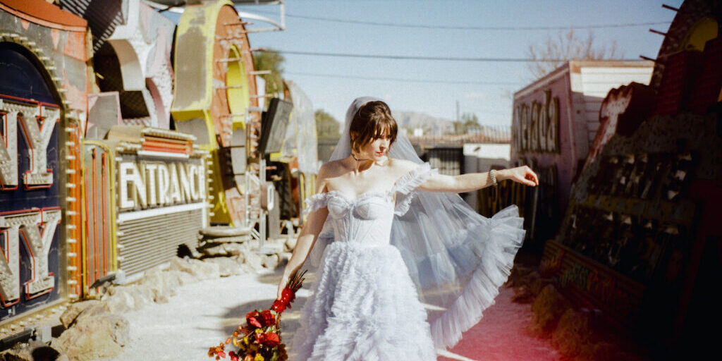 A bride in a white ruffled dress and veil stands outdoors holding a bouquet of red flowers, beside vintage neon signs under a clear sky.