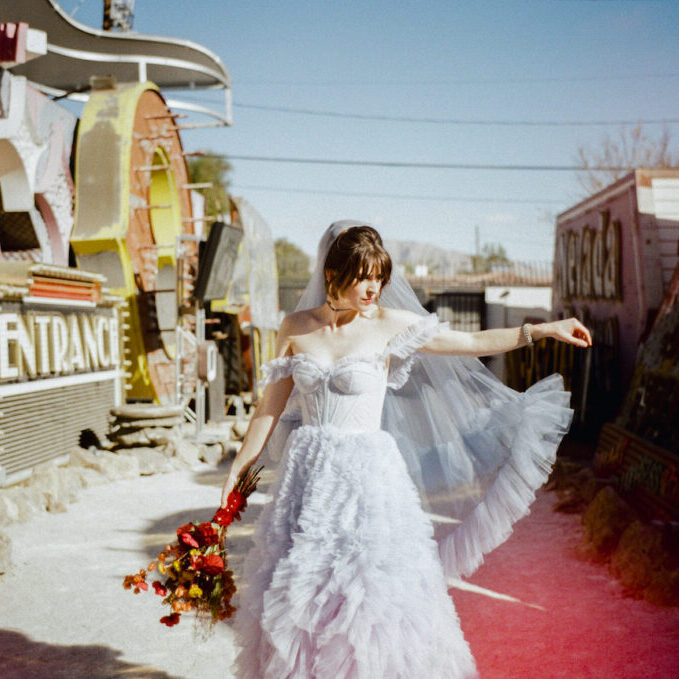 A bride in a white ruffled dress and veil stands outdoors holding a bouquet of red flowers, beside vintage neon signs under a clear sky.