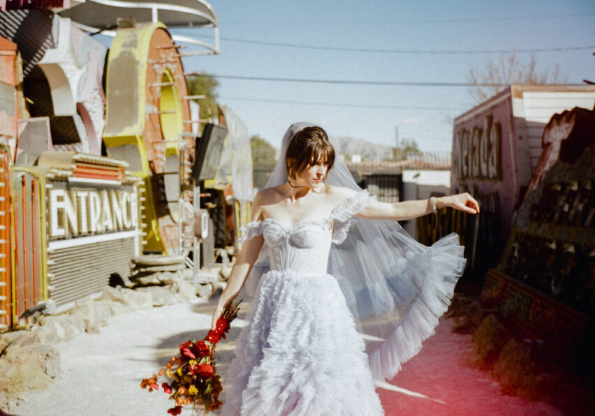 A bride in a white ruffled dress and veil stands outdoors holding a bouquet of red flowers, beside vintage neon signs under a clear sky.