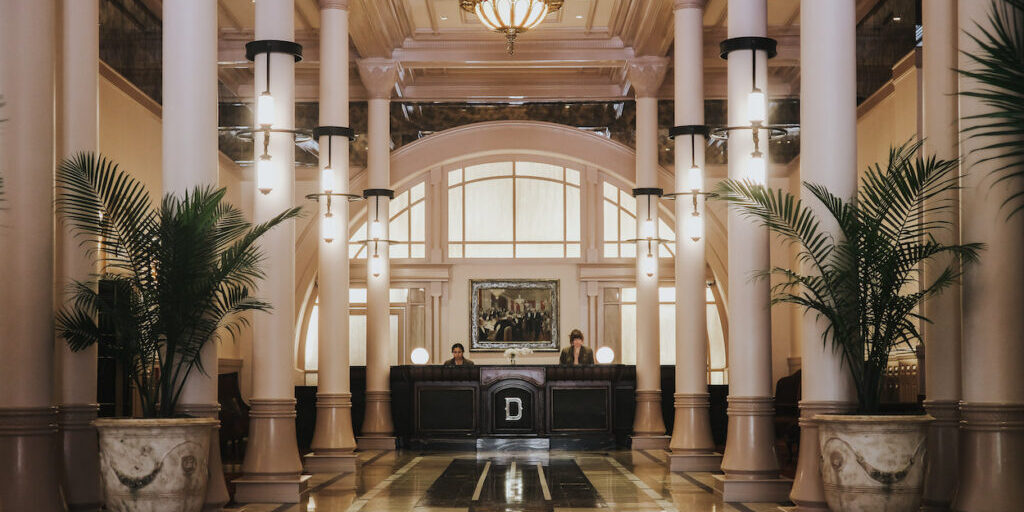 Hotel lobby with marble floors, tall white columns, a chandelier, two palm plants, and two people standing behind a black reception desk.