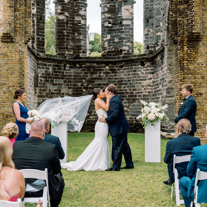 A bride and groom share a kiss at the altar under a flowing veil within the ruins of an ancient stone structure, as the bridal party and guests look on. Bridesmaids in blue dresses hold white bouquets, adding a touch of elegance to the rustic setting.