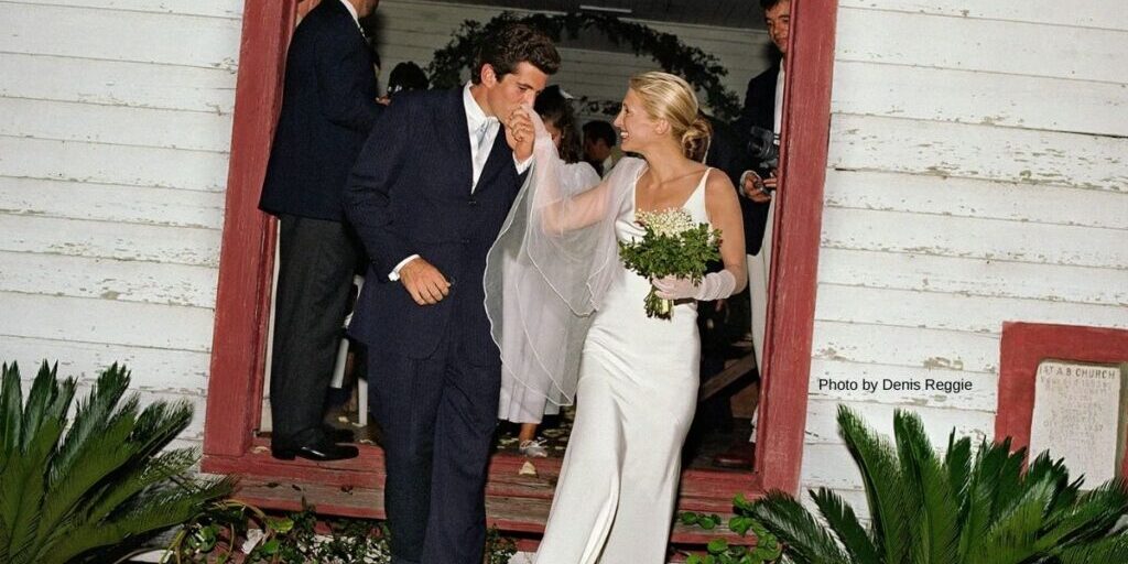 A groom kisses the hand of his bride as they exit a building; she wears a white dress and holds a bouquet, while guests stand in the background.