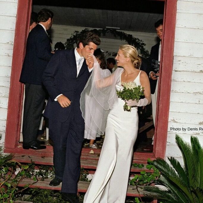 A groom kisses the hand of his bride as they exit a building; she wears a white dress and holds a bouquet, while guests stand in the background.