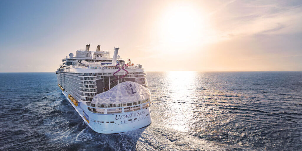 A large cruise ship sails on the ocean under a clear sky with the sun setting on the horizon.