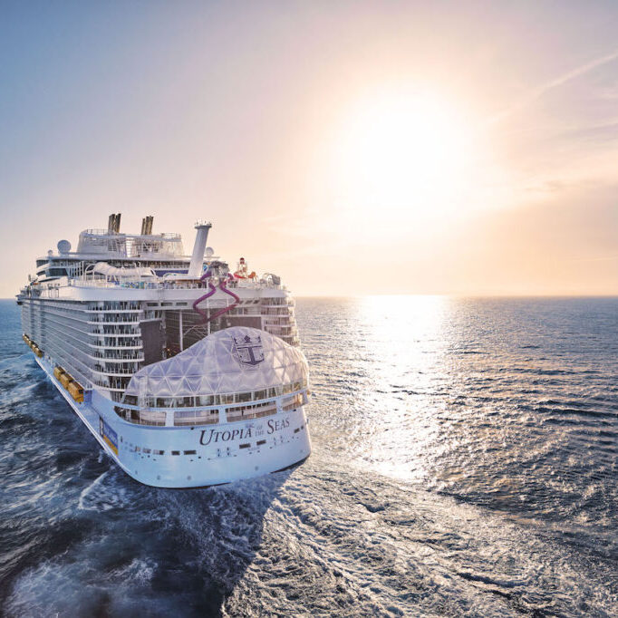 A large cruise ship sails on the ocean under a clear sky with the sun setting on the horizon.