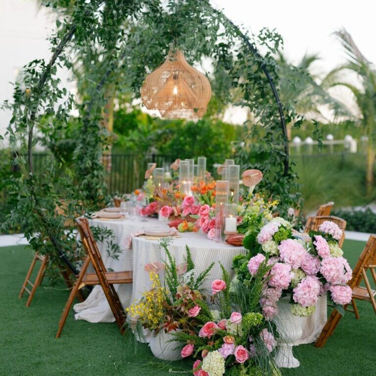 A round outdoor table with a white tablecloth is decorated with pink flowers and greenery, surrounded by wooden chairs and set under a leafy arch with a hanging woven light fixture.