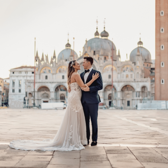 Bride and groom stand together in front of a historic building with domes and arches. The bride wears a long white gown, and the groom is in a dark suit.