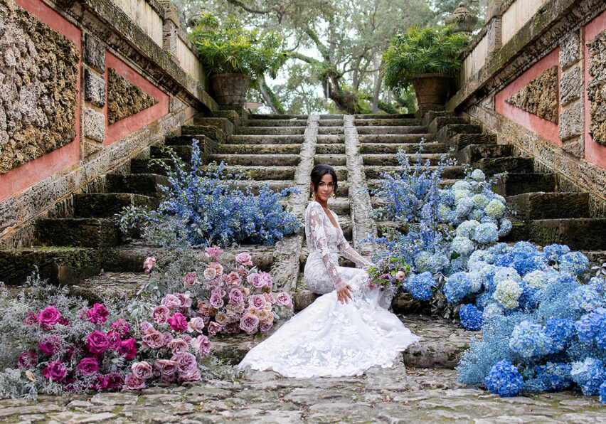 bride sits at bottom of stairs surrounded by cloud-like blue and pink floral arrangements