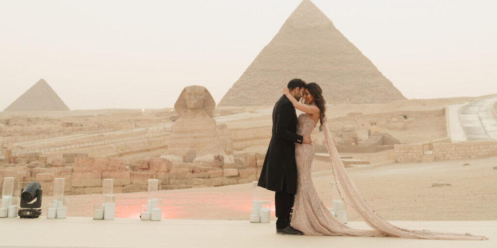 A couple dressed formally embraces on a platform near the Sphinx and a pyramid in Egypt, with soft lighting and candles arranged along the edge.