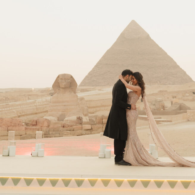A couple dressed formally embraces on a platform near the Sphinx and a pyramid in Egypt, with soft lighting and candles arranged along the edge.
