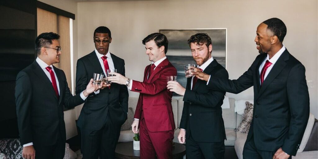 Five men in suits, one in a red suit and four in black suits, stand in a living room toasting with glasses.