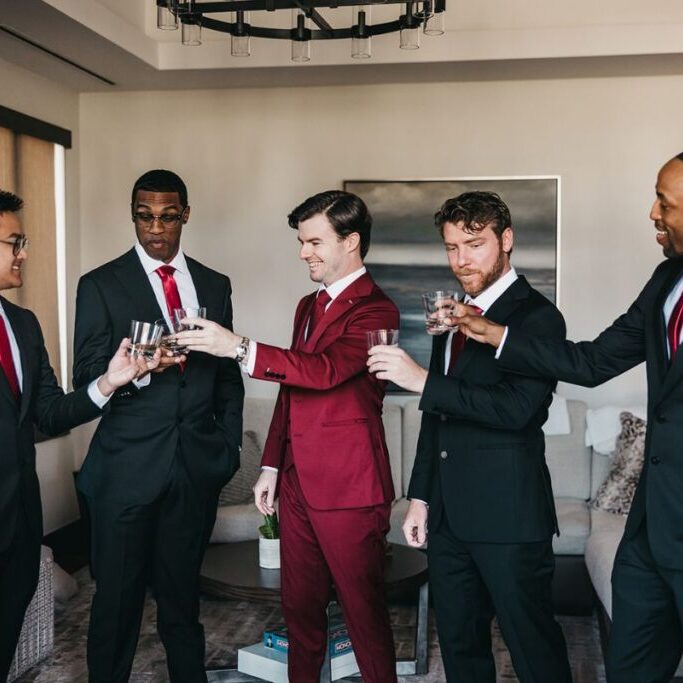 Five men in suits, one in a red suit and four in black suits, stand in a living room toasting with glasses.