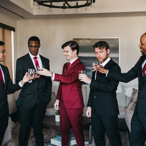 Five men in suits, one in a red suit and four in black suits, stand in a living room toasting with glasses.