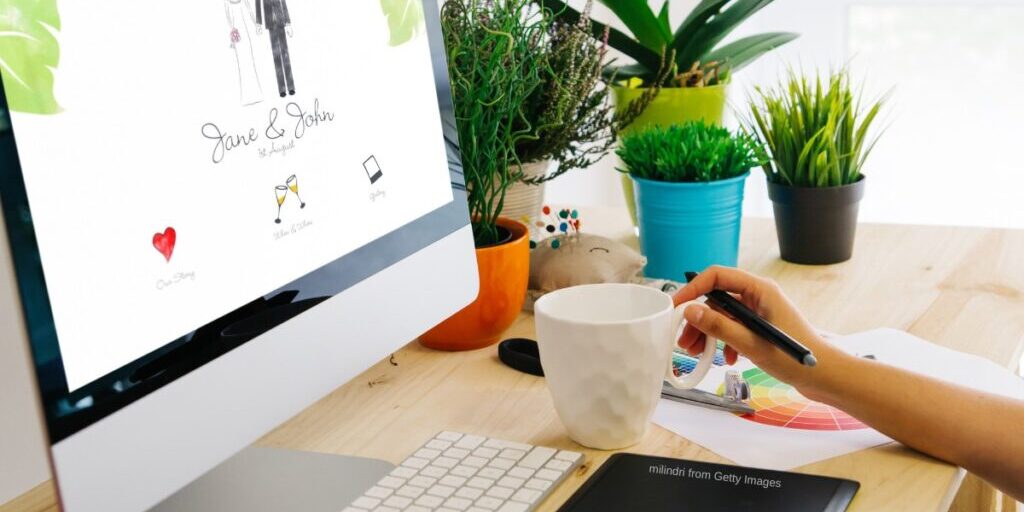 person sits at desk looking at wedding website on large desktop screen with coffee and plants sitting on the desk to the right